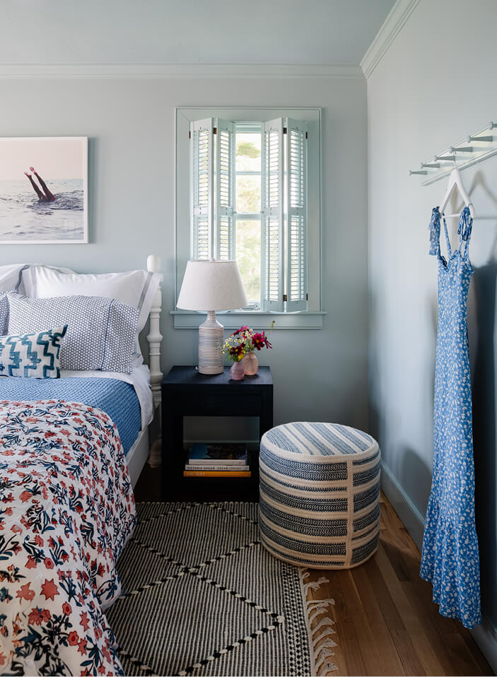 Light blue guest bedroom with striped rug, floral bedding, louvered shades, and playful art in Martha’s Vineyard by Cape Cod interior designers.