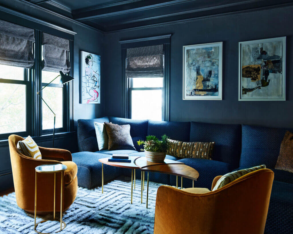 Navy blue media room design in a historic Brookline home with dark walls and a matching coffered ceiling. A modern sectional blends with navy walls, reupholstered swivel chairs, and personal art.