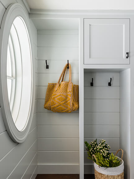 Light grey custom cabinets with inset details, traditional door style, and shiplap, featuring an oval window in a New England traditional mudroom.