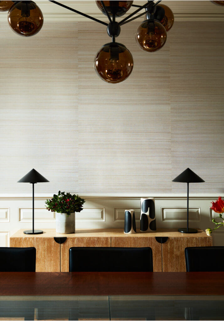 A dining room with grasscloth wallcovering, a modern console, Roll & Hill light fixture, and a custom walnut and glass dining table, complemented by black lamps.