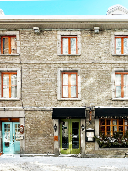 Visit Montreal in the Winter, showcasing a historic stone facade with natural wood windows, lime green and aqua painted doors, fresh snow, and a bright blue sky.