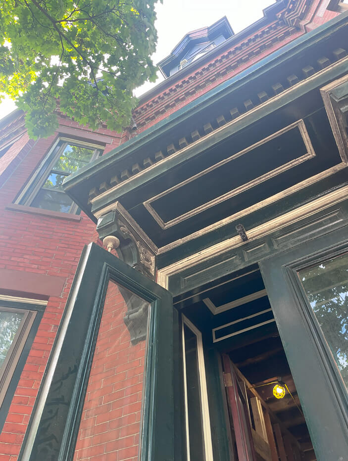 A close-up of the brownstone's brick exterior, green pediment, intricate corbels with dentil molding, and original green doors with vintage hardware.