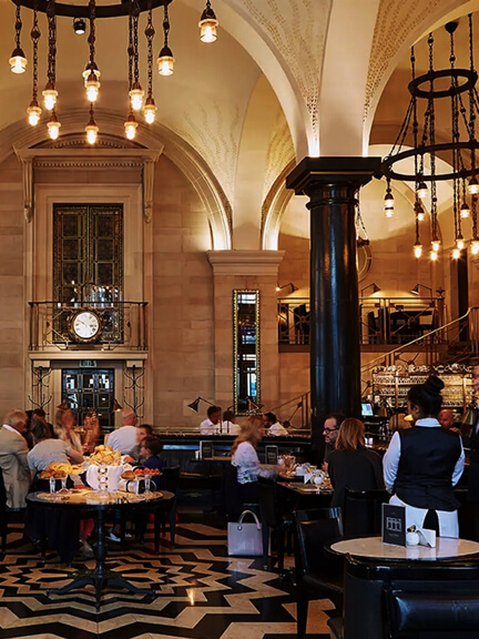 London restaurant design at The Wolseley features warm stone walls, black and white patterned flooring, and grand iron chandeliers in a classic cafe interior.
