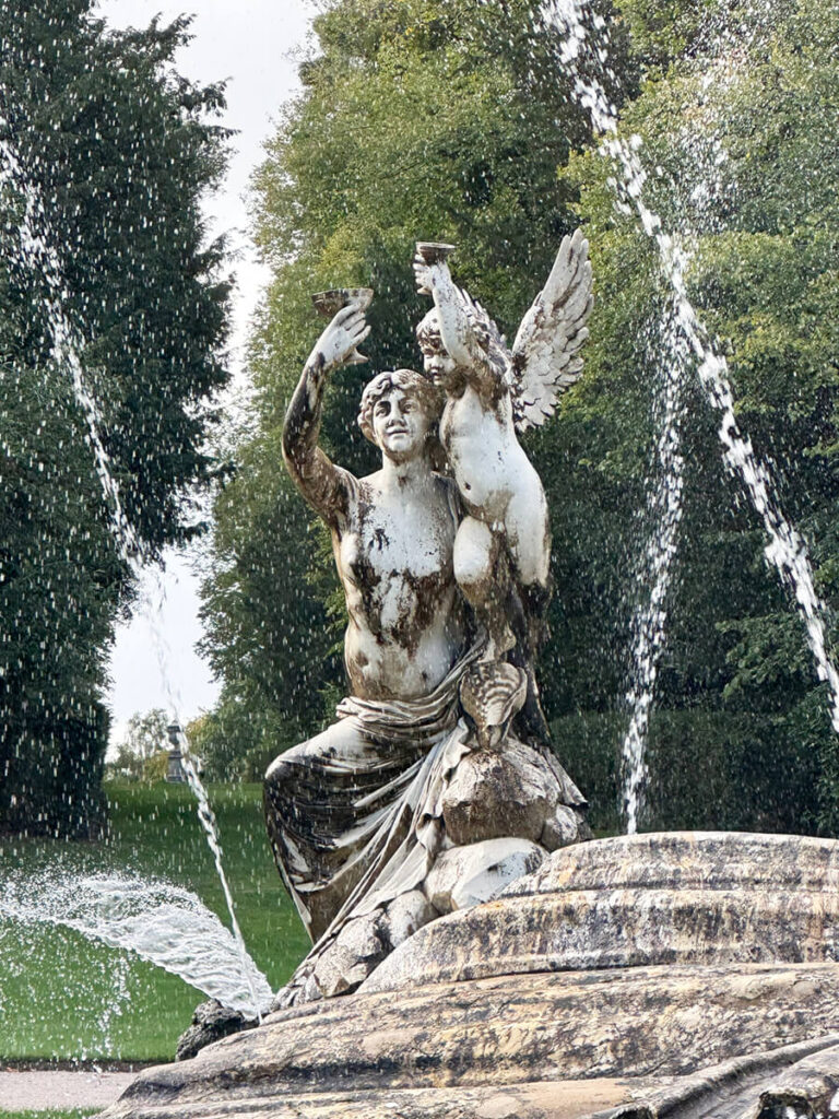 Sculptural fountain at the entry of Cliveden House, featuring carved stone figures, cascading water, and formal garden surroundings.