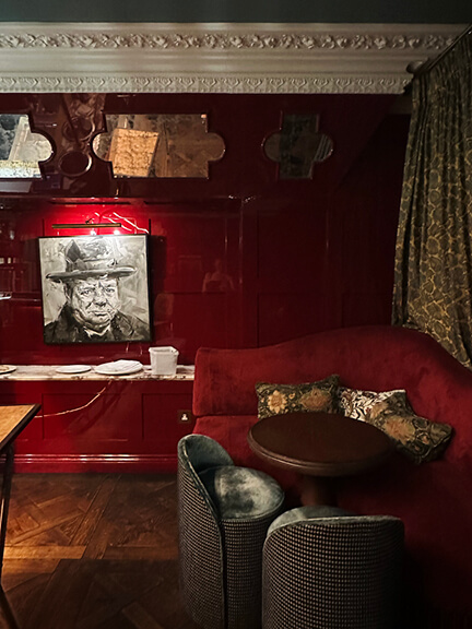 Glossy red walls, red velvet furniture, bold art, and floral drapery panels at the bar in No 50 Cheyne with grand historic molding.