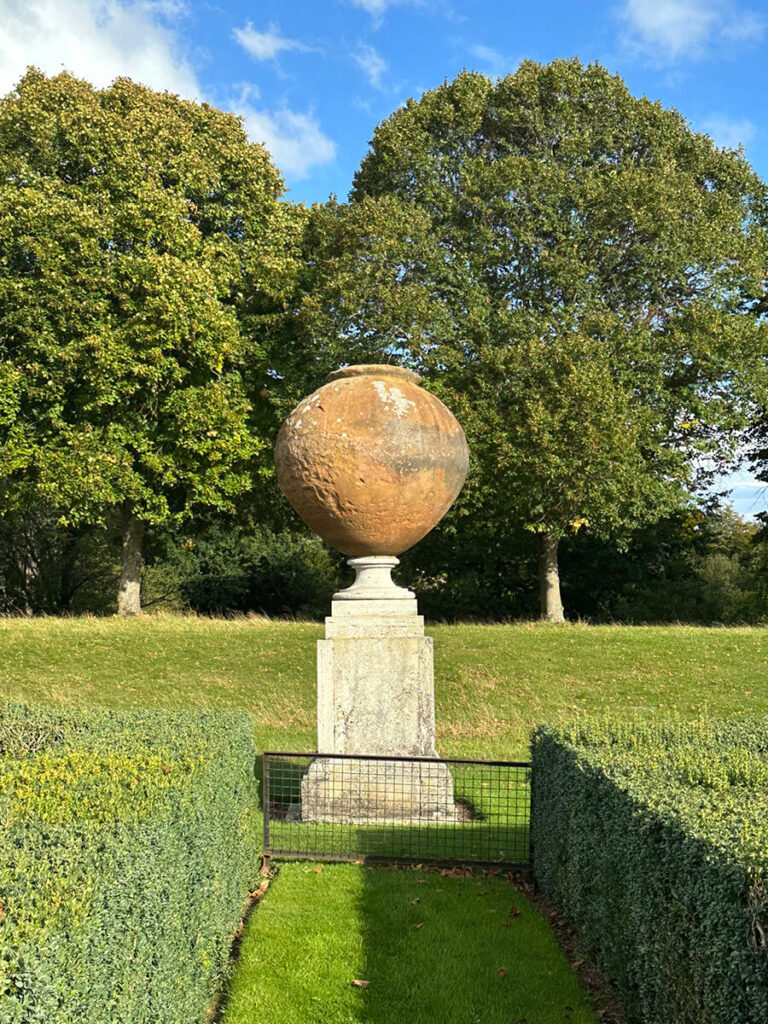 Garden urn in a classic English countryside setting, surrounded by manicured hedges, stone balustrades, and seasonal plantings.