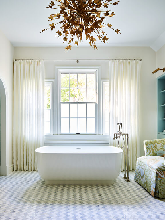 Freestanding tub on leaf-shaped marble mosaic floor with embroidered linen drapery, brass gilded light fixtures, blue cabinets, and a Christopher Farr slipper chair