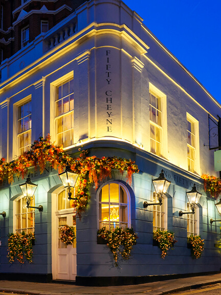 Night view of No 50 Cheyne in London with glowing lighting, blue and white historic facade, and floral details on the corner building.