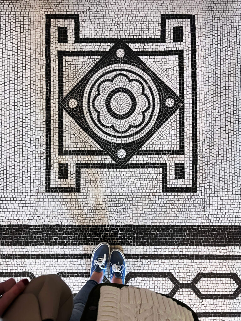 Victorian tile patterns appear in a black and white geometric mosaic floor with circles and concentric squares inside a historic London museum.