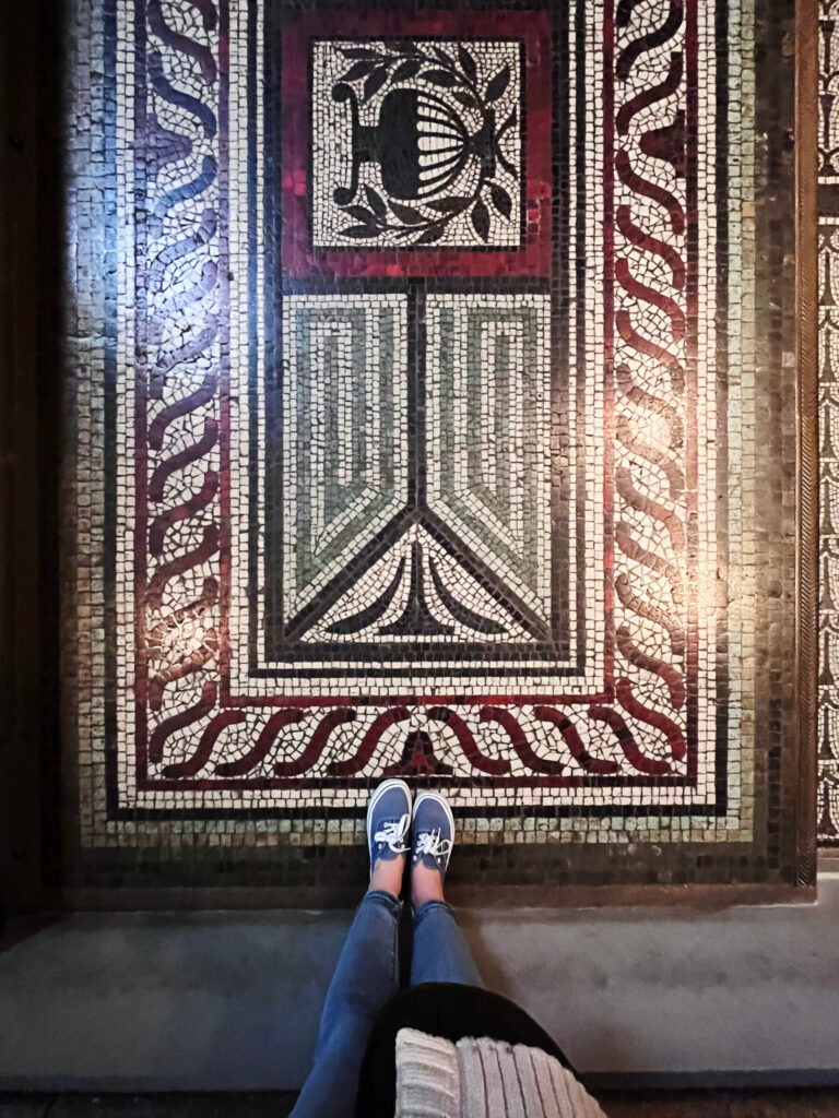 Victorian tile patterns appear in a red, green, and black mosaic floor with Grecian-inspired motifs inside a historic building in London.