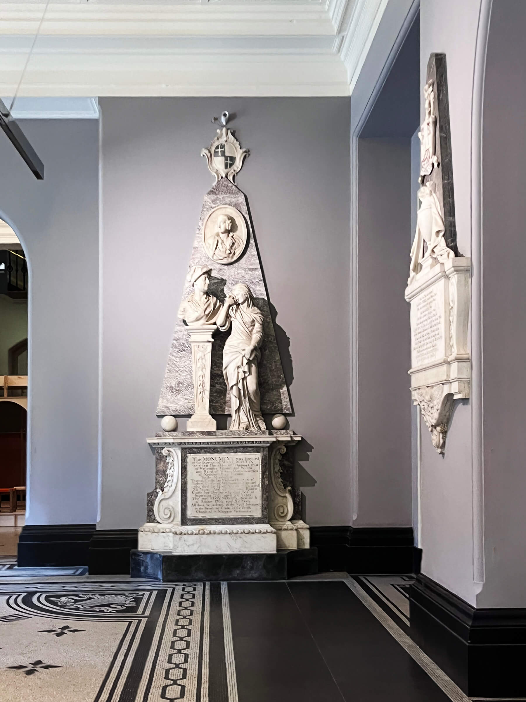 A Grecian sculpture stands beside black and white mosaic tile flooring with a Greek fret border inside the Victoria and Albert Museum in London.
