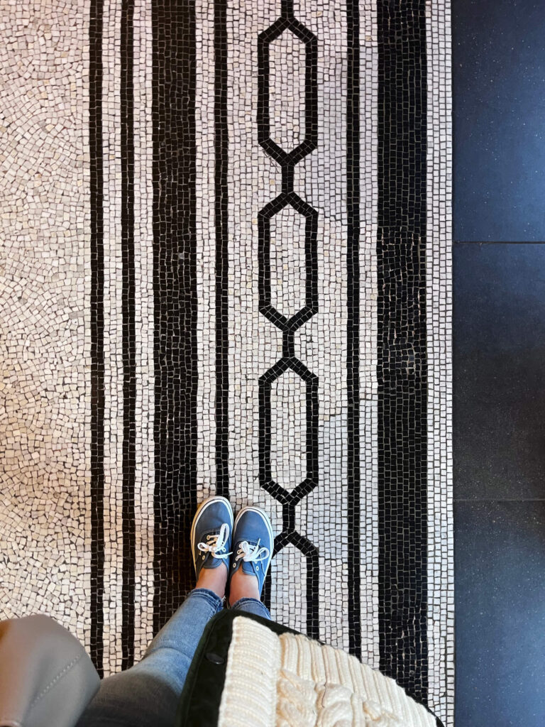 Victorian tile patterns appear in black and white Greek fret mosaic flooring inside a historic London museum with European architectural influence.