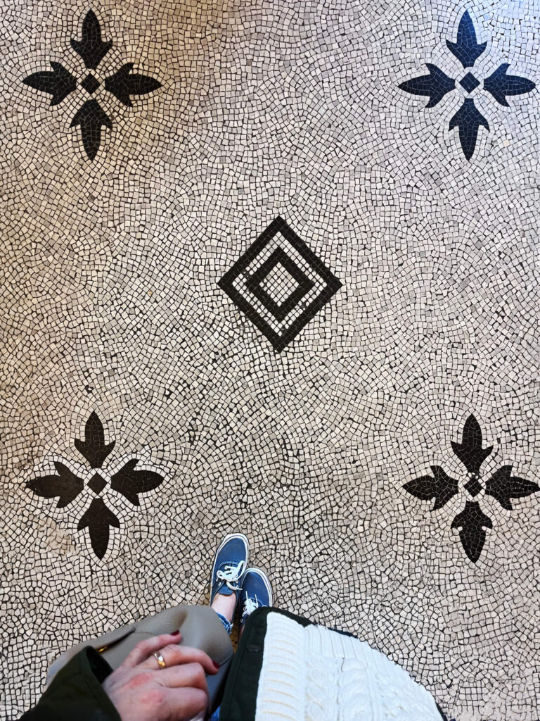 Victorian tile patterns appear in white and black mosaic flooring with geometric shapes and subtle fleur-de-lis motifs inside a London museum.