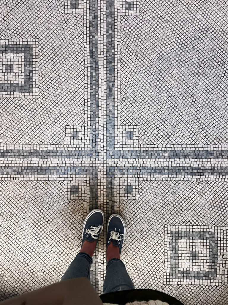 Victorian tile patterns appear in white and black mosaic flooring with geometric shapes and subtle fleur-de-lis motifs inside a London museum.