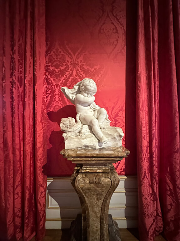 Kensington Palace Interiors featuring a marble cherub statue set against red damask walls in a historic room.