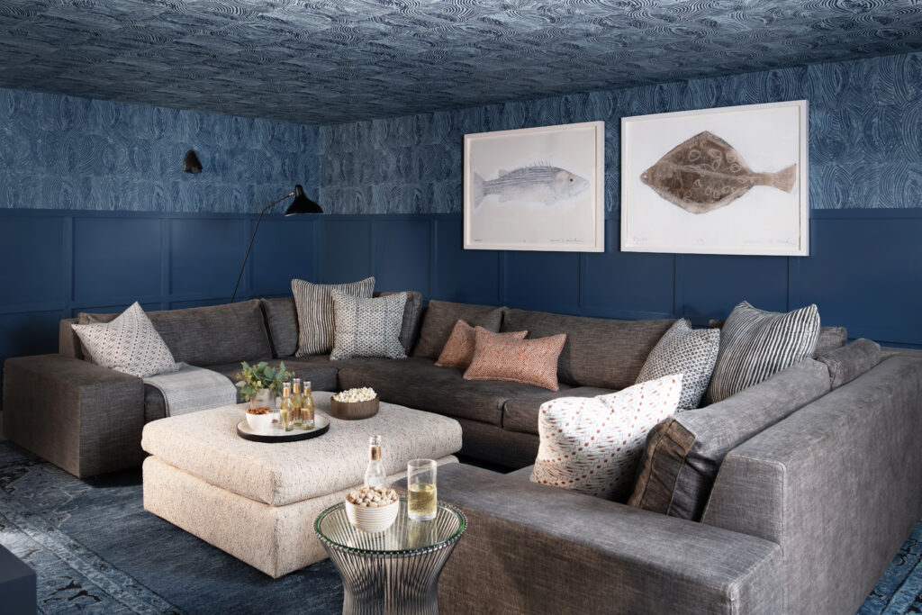 Dark and moody basement media room in Hingham designed by Sarah Scales, with dark millwork, wallpapered walls and ceiling, charcoal sectional, custom ottoman, and navy blue tones creating a cozy space without natural light.