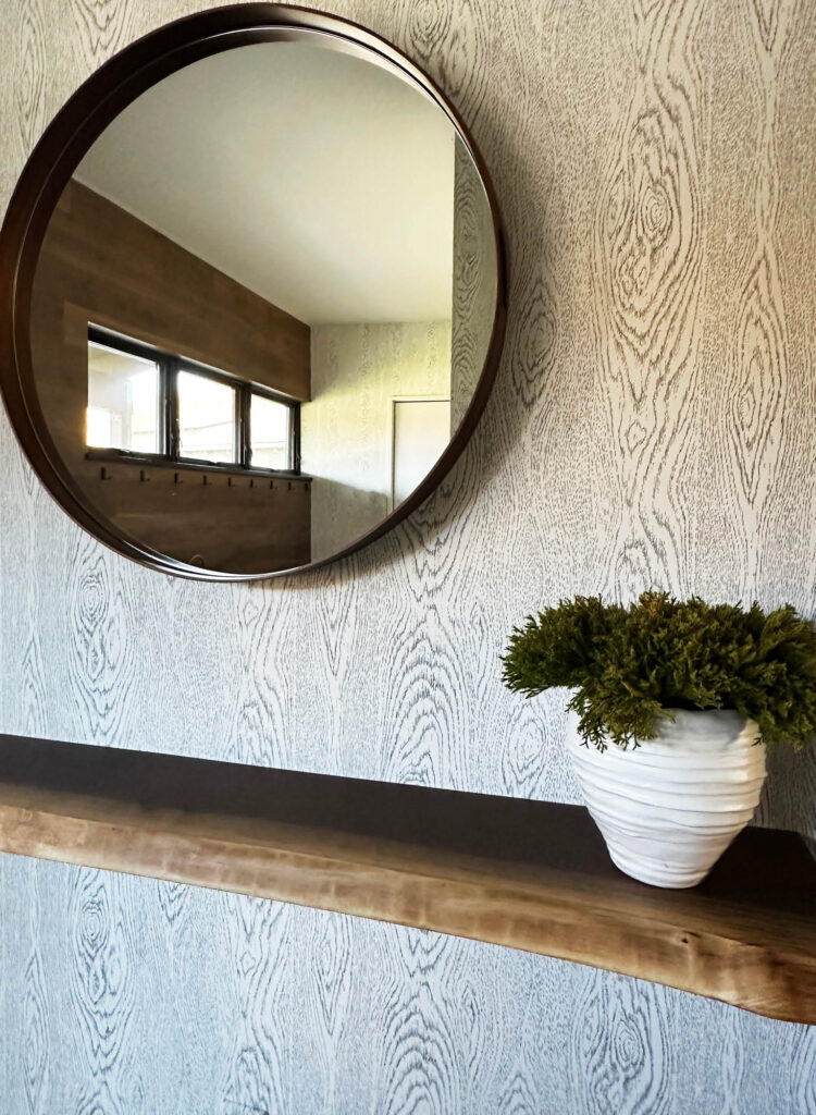 Foyer with faux bois wallpaper, walnut surface, and round wood mirror in Vermont interior design project.