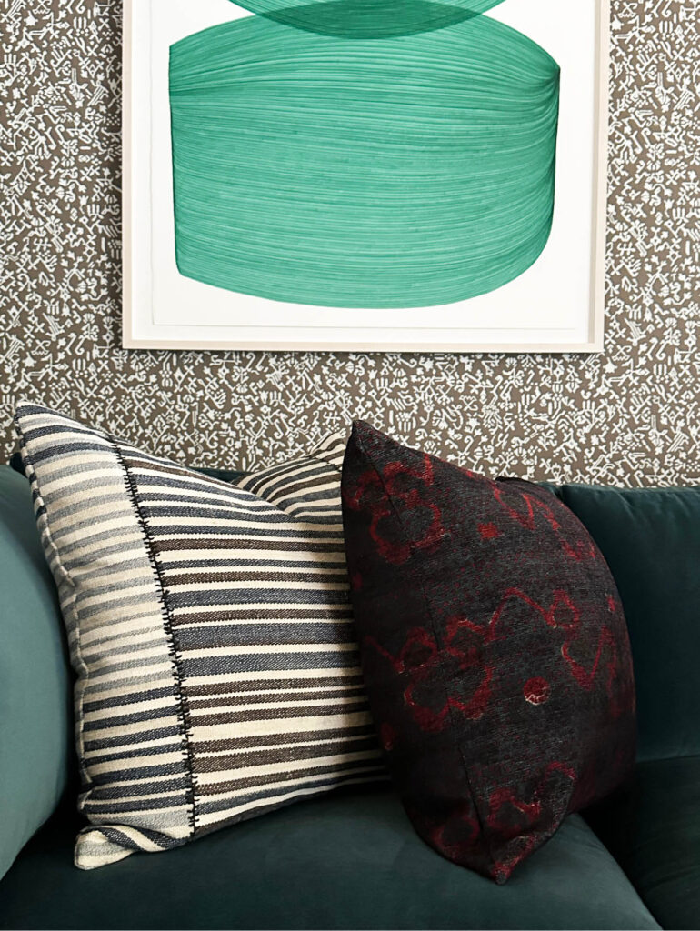 Custom pillows on a teal velvet sofa with brown and white patterned wallpaper and bold green art in a Vermont mountain home.