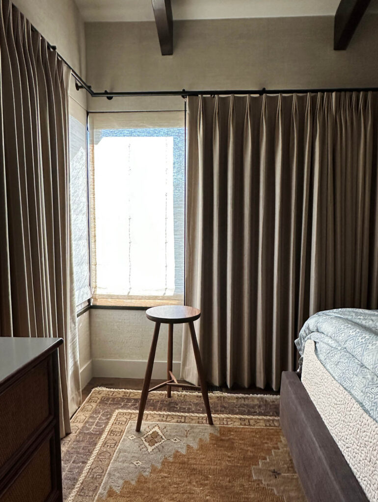 Primary bedroom with vintage-inspired rug, custom camel wool drapery, matching Roman shades, grasscloth wallpaper, and a purple linen bed frame in a Vermont mountain home.