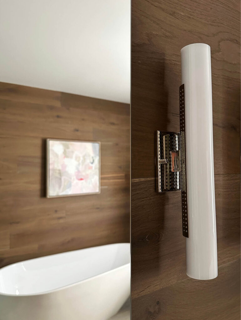 Polished nickel sconces and custom art above the freestanding tub in the primary bathroom of a Vermont mountain home.