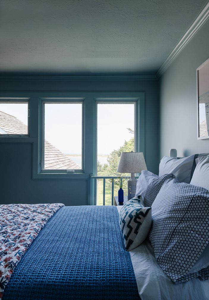 Guest bedroom with soft blue walls and ceiling, a waterfront view of the bay, simple floral bedding, and curated artwork. Designed by Boston interior designer Sarah Scales, this space captures coastal and New England interior design, perfect for guests.