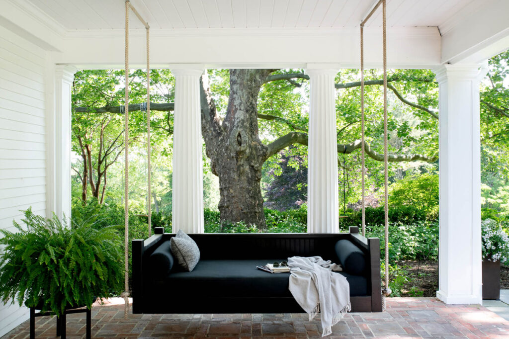 Hanging black daybed on a covered porch with Greek columns and brick pavers, overlooking a mature garden at a historic Concord home.