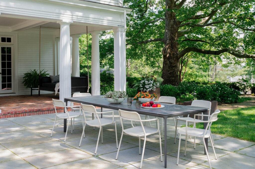 Covered porch with Greek columns, daybed, bluestone patio, dining table, and views of a mature garden at a historic Concord home.