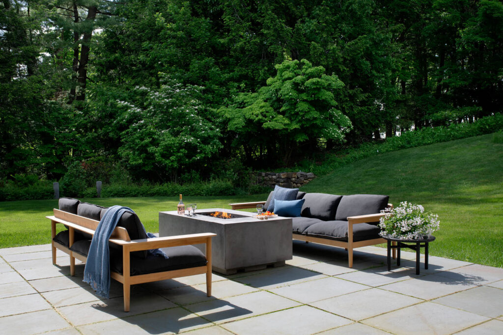 Bluestone patio with black and teak-facing sofas, a central gas fireplace, and views of a mature garden at a historic Concord home.