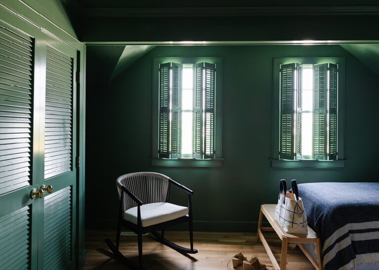 Dark green guest bedroom with louvered doors, window shades, modern rocking chair, and cane bench in Edgartown by a Cape Cod interior designer.