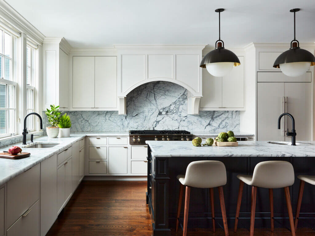 A classic white kitchen with simple cabinet doors and subtle edge pulls, a black island, black metal fixtures, and modern leather stools. 