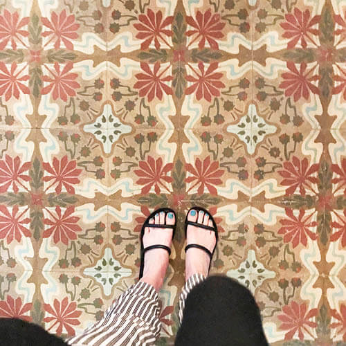 Historic concrete tiles in a Havana, Cuba home with red, brown, and light blue patterns. Sarah Scales in striped pants and black sandals.