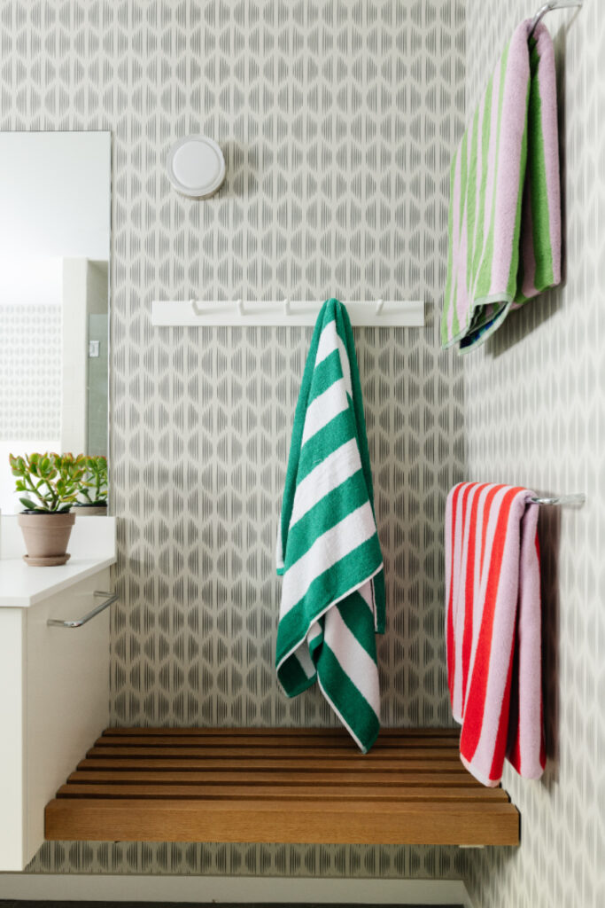 Kid-friendly bathroom with Schumacher wallpaper, a white vanity, teak bench, and colorful striped towels in a basement bathroom.