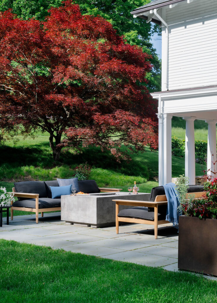 Classic architecture with Greek columns, white siding, and a bluestone patio with teak and black sofas, gas firepit, and red maple tree.