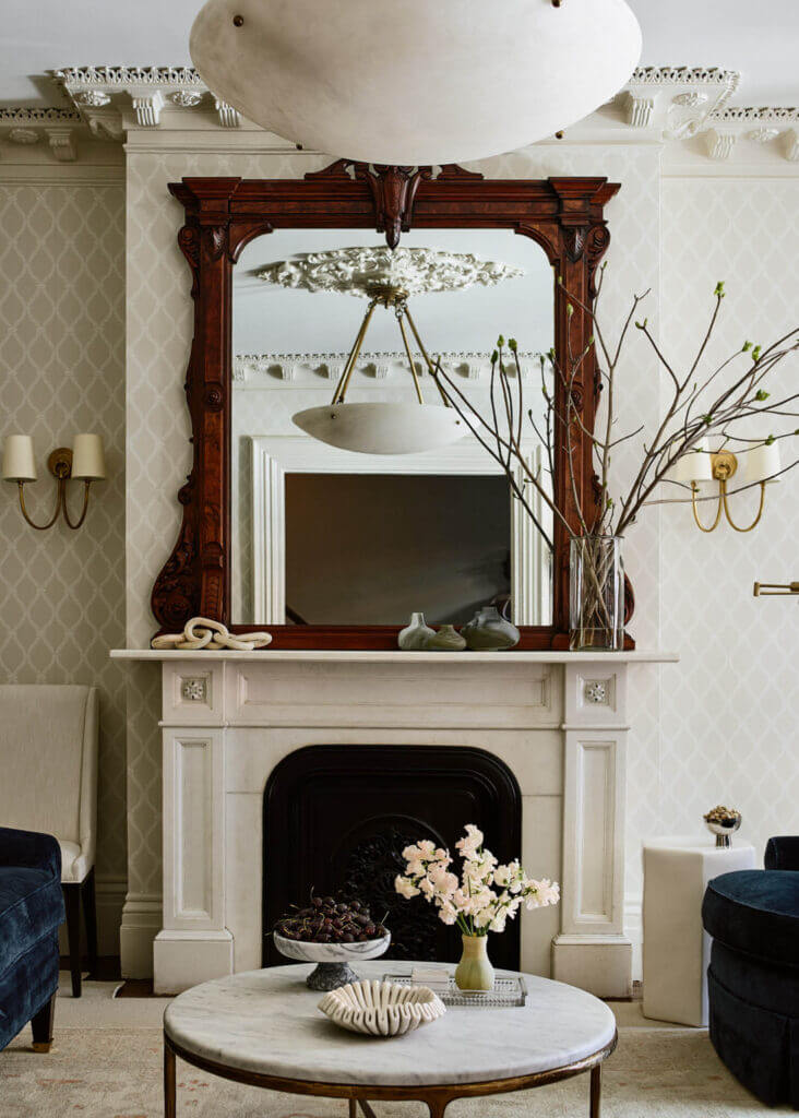 A historic brownstone’s original marble fireplace with a wood mirror, trellis wallpaper, and brass sconces reflects classic historic home fireplace design.