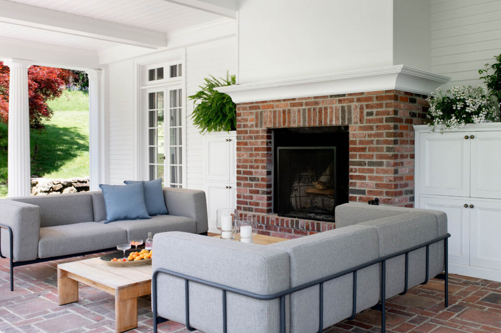 Covered porch with Greek columns, brick pavers, a wood-burning fireplace, and modern furniture by Boston interior design firms.