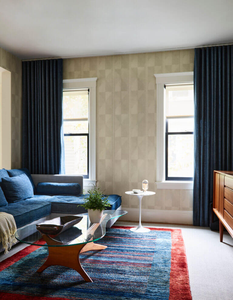 A reupholstered Ligne Roset sectional in denim and grey, with custom drapery, Pierre Frey wallpaper, and a vintage rug in bold red and blue.