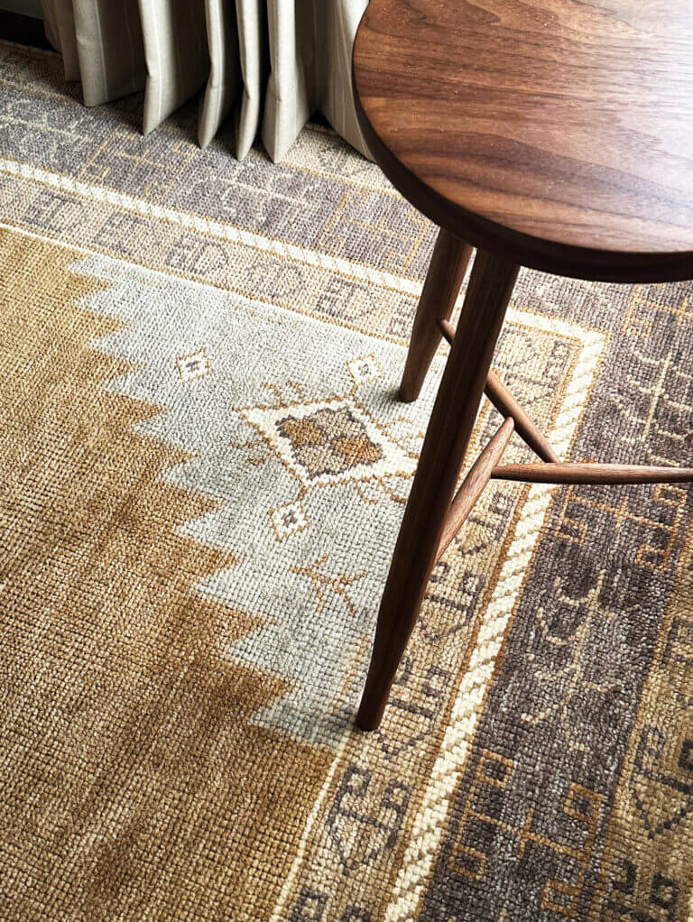 A handmade walnut stool, camel wool drapery panels, and a vintage-inspired area rug in a Vermont ski house. Designed by Stowe interior decorators.