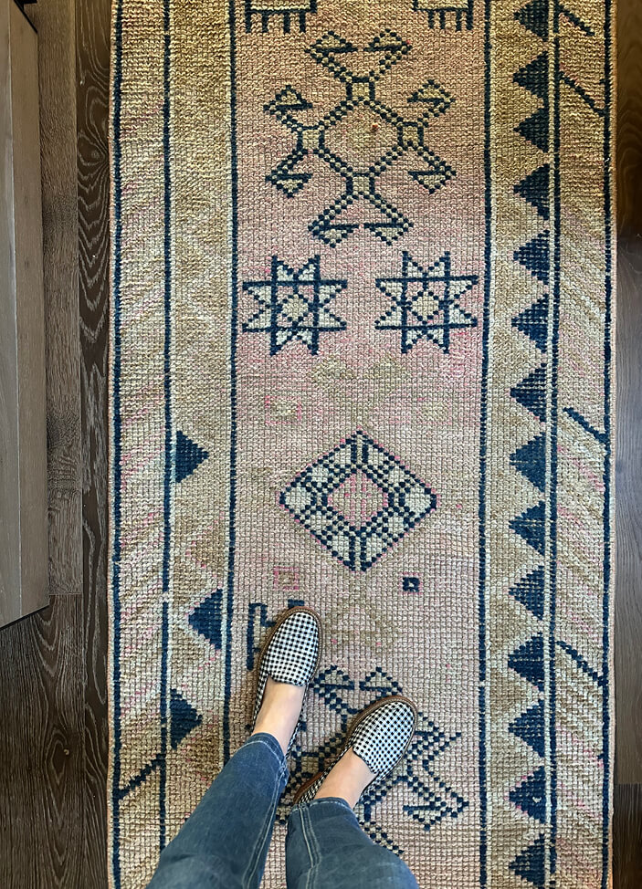 A vintage area rug in beige, soft pink, and navy blue on a dark wood floor in a Vermont ski house, styled with navy and white gingham loafers.