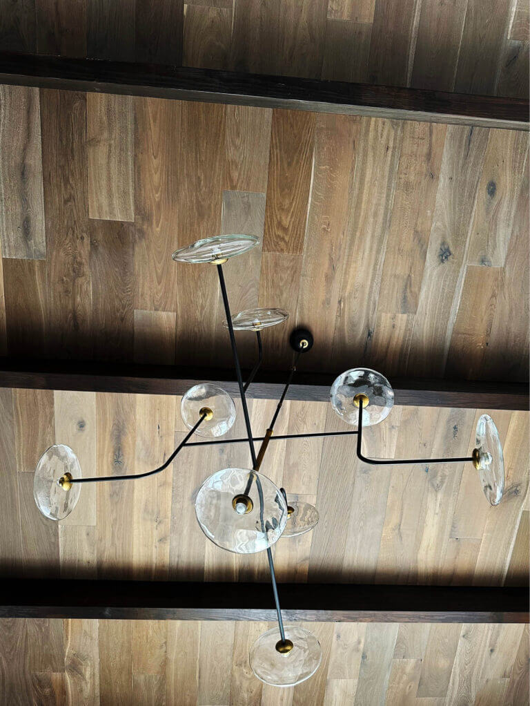 A Visual Comforts chandelier with glass, black metal, and brass accents hangs from a wood ceiling with exposed beams in a Vermont ski house living room.