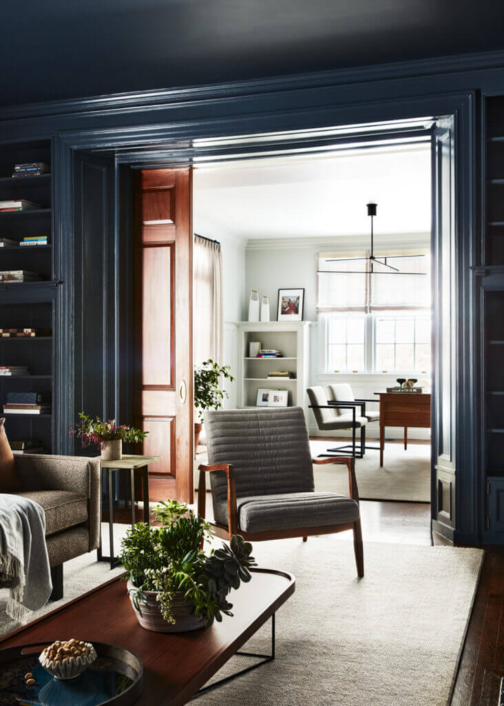 A navy media room with original millwork, built-ins, and natural wood doors, opening to a light grey office with mid-century inspired furniture and neutral spaces.