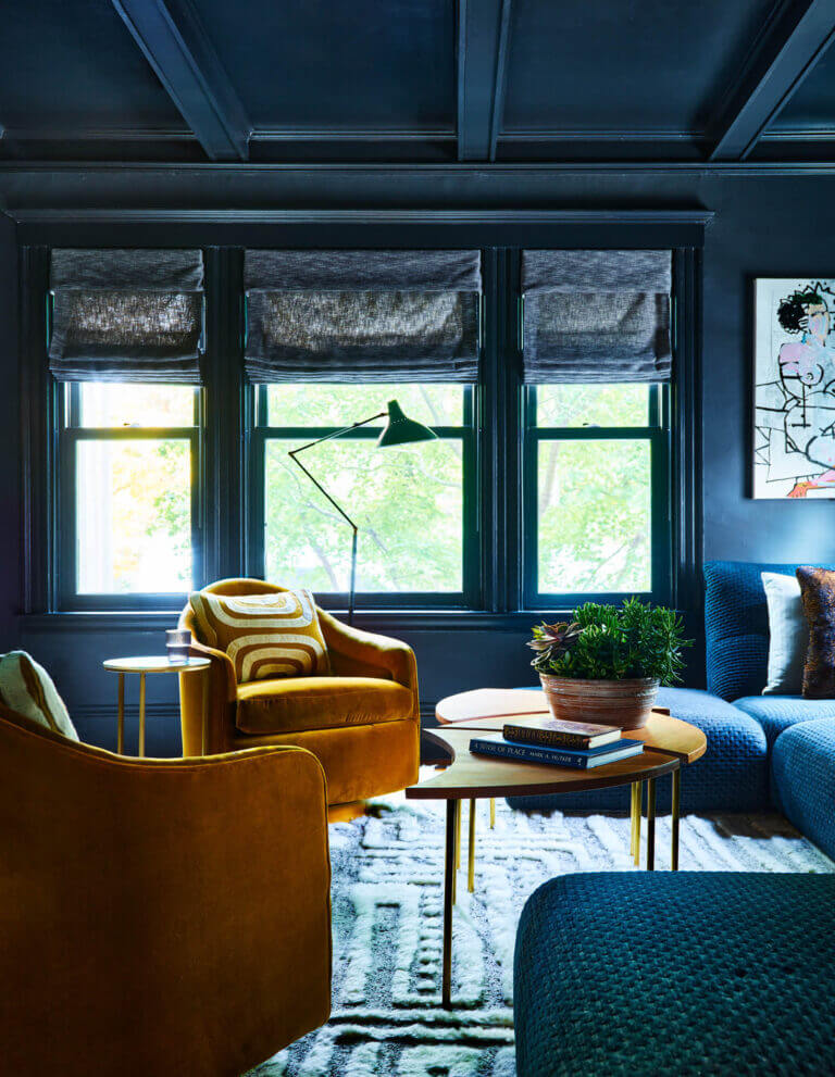 A deep navy lounge space with a coffered ceiling, modern sectional, reupholstered swivel chairs, vintage coffee table, and dark grey Roman shades.