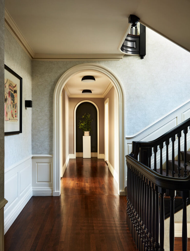 A second-floor corridor with original millwork, black-painted stairs, geometric wallpaper, modern black light fixtures, and colorful art in a historic Concord estate.