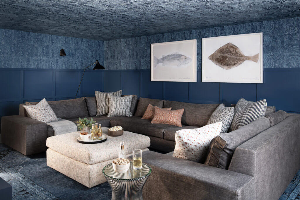 Navy blue media room design in a new Hingham home with navy wainscot, blue and green patterned wallpaper, a modern grey sectional, and a light ottoman.