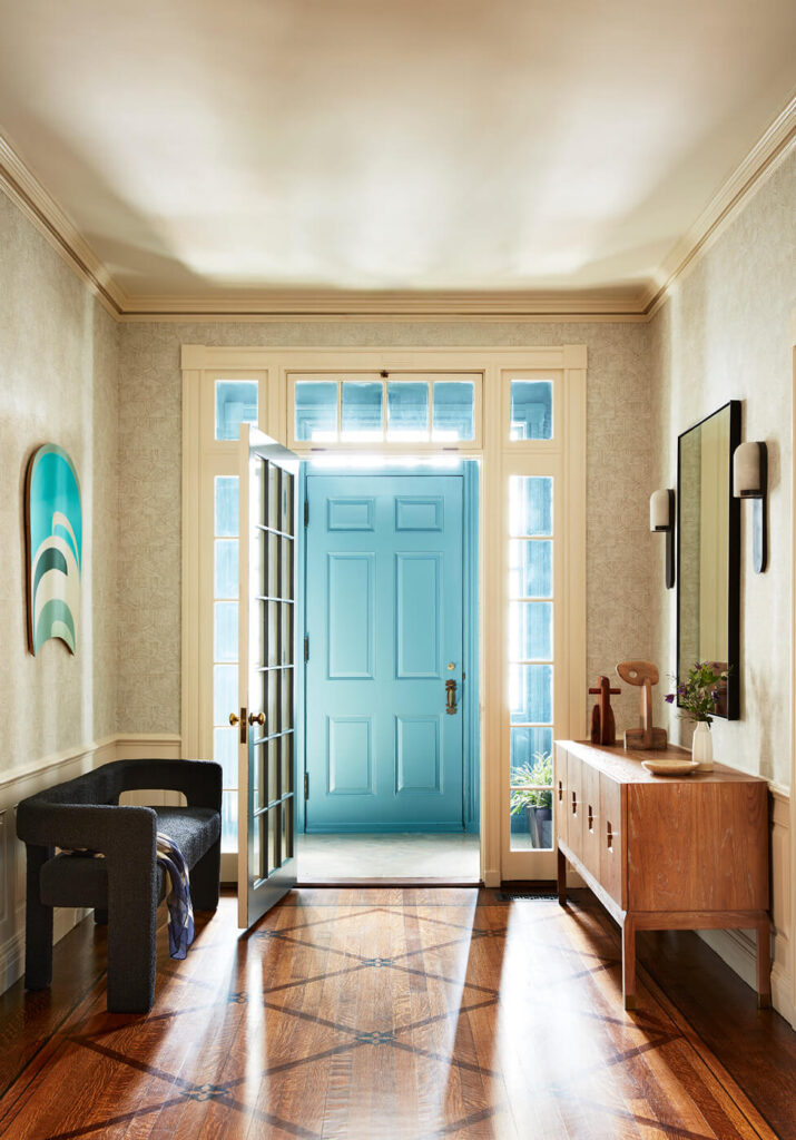 A formal foyer with sensicle wood flooring, off-white wainscoting, geometric wallpaper, fun art, and modern furniture, looking through to a small blue entry.