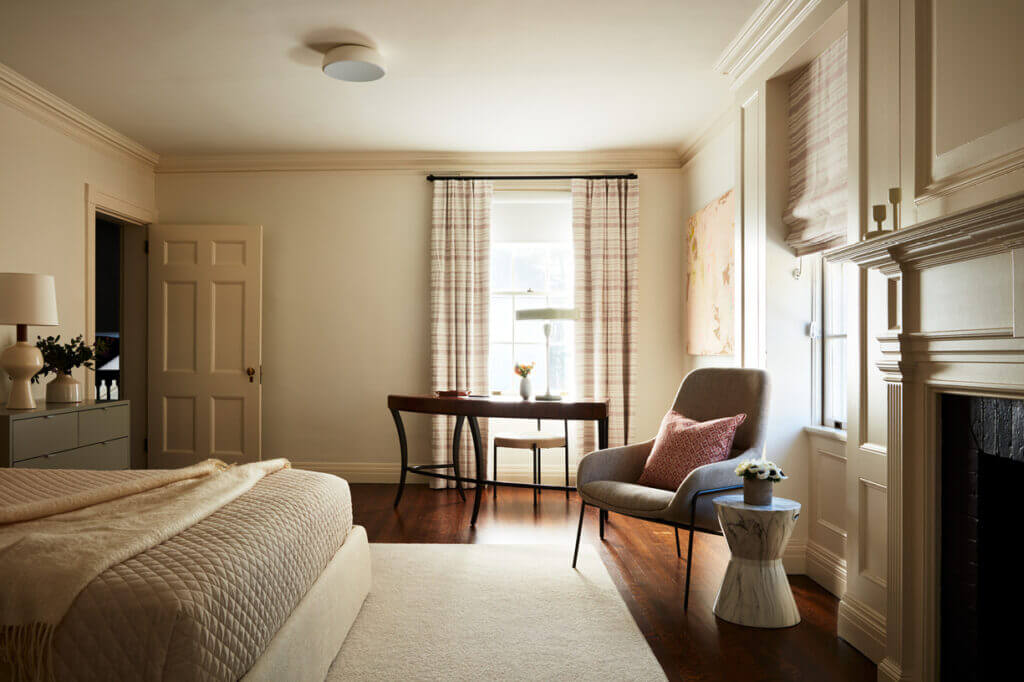 Historic Home Interior Design in a primary bedroom with a king bed, neutral area rug, off-white walls, and an original fireplace, mixed with modern and vintage furniture.