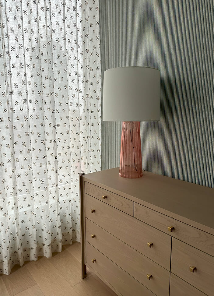 A primary bedroom with light green and blue Zak and Fox wallpaper, light oak dresser from Made Goods, and a pink glass lamp with a white shade.