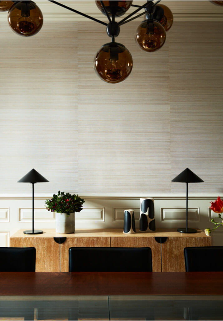 A dining room with grasscloth wallcovering, a modern console, Roll & Hill light fixture, and a custom walnut and glass dining table, complemented by black lamps.