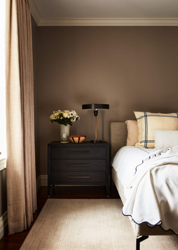 A primary bedroom with a simple upholstered bed, dark oak bedside tables, beige linen drapery panels, and a Visual Comfort lamp.