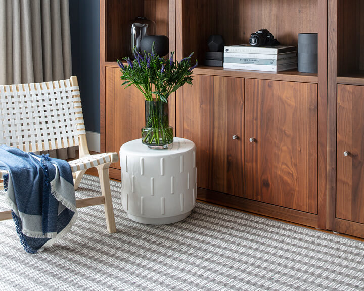 A custom-cut area rug in a primary bedroom of a new construction home, featuring walnut bookshelves, a white leather chair, and a ceramic side table.