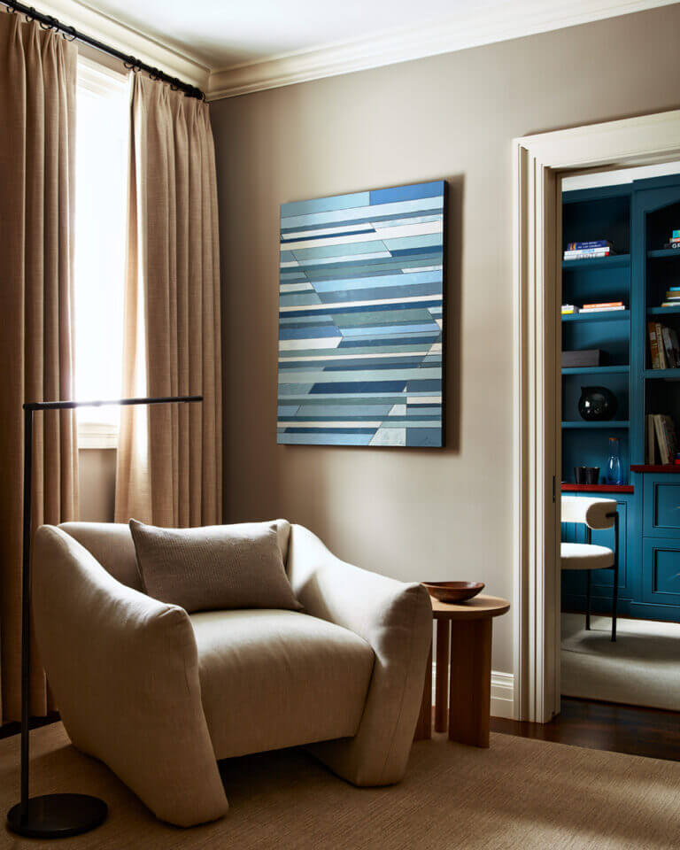 A primary suite sitting room with an oversized chair from Maiden Home, sleek floor lamp from Visual Comforts, oak side table, and blue art, looking into the navy blue office.